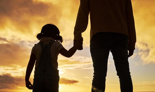 A shadowed, rear-view image of a man and child holding hands looking out against a sunset sky.
