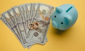 A top-down shot of two wedding rings resting on a fan of bank notes next to a light cyan piggy bank.