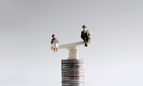 A miniature man and woman with a baby, seated on a seesaw resting on a stack of coins, weighted toward the woman's side.