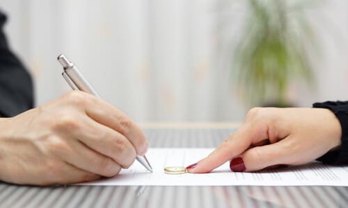 A woman's hand sliding her wedding ring across a settlement agreement being signed by a man's hand.