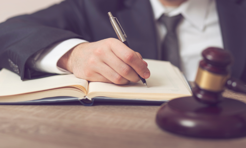 A lawyer drafting out an estate plan in a large notebook on his desk with an out of focus gavel in the foreground.