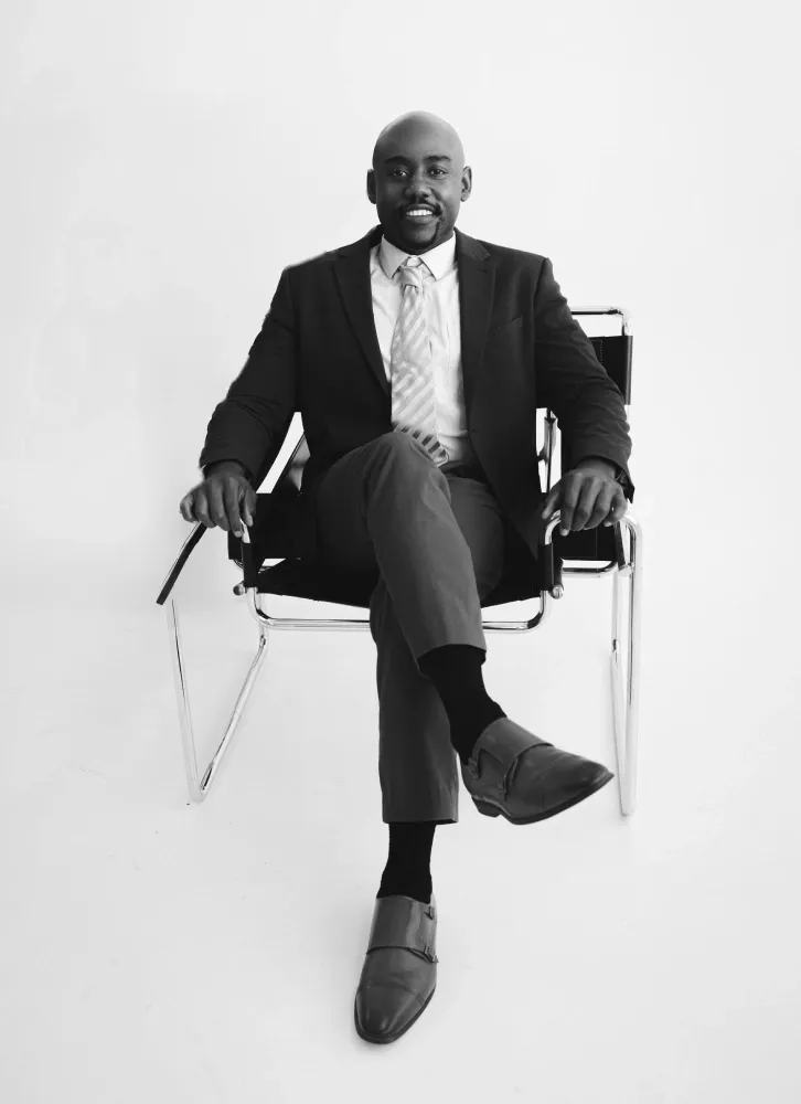 Alex Sanders seated in a chair wearing a suit and tie, presented in a timeless portrait style.